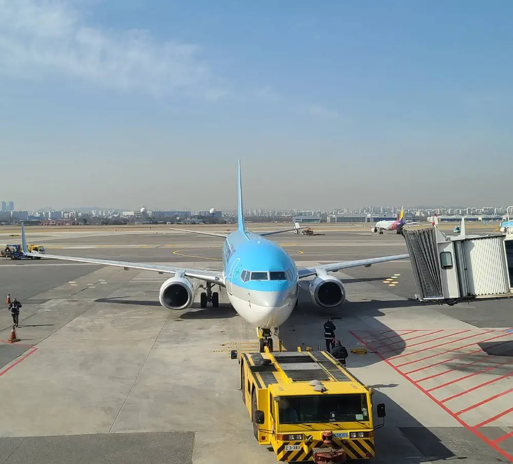 A light blue Korean Air airplane parked on the airport runway connected to a boarding jet bridge