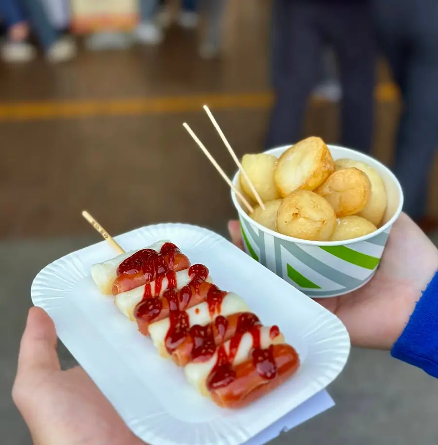 A person holding a skewer of Sotteok-Sotteok with red sauce and a cup of roasted potatoes at a Korean highway rest stop