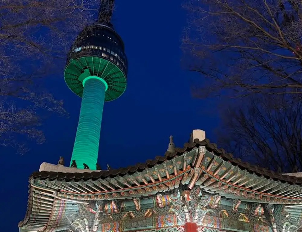 N Seoul Tower illuminated in green at night behind a traditional Korean pavilion roof under a dark blue sky